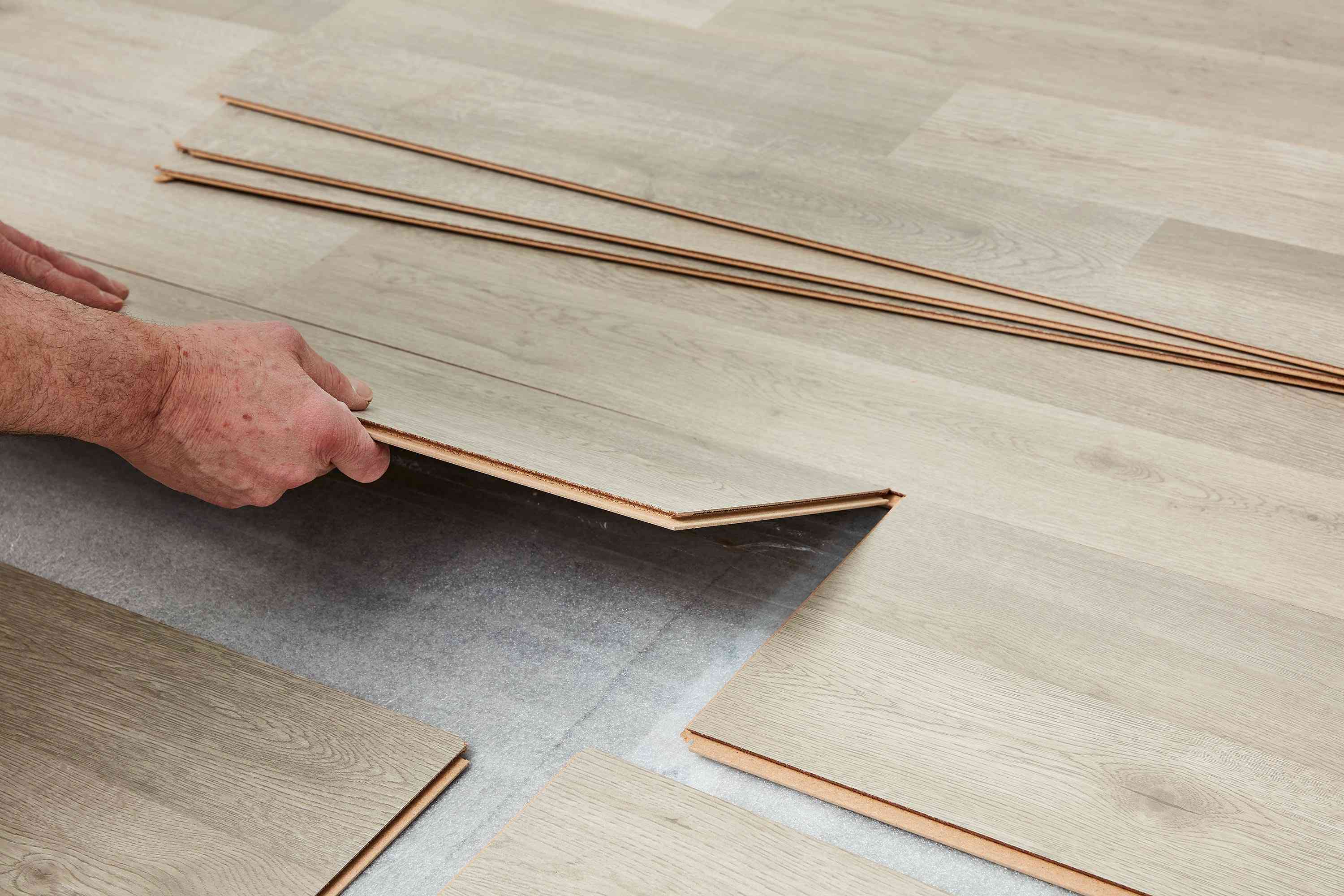 Wide practical photo of hands installing light wood-toned laminate flooring planks, neutral beige and tan tones, methodical and calm mood, shallow depth of field