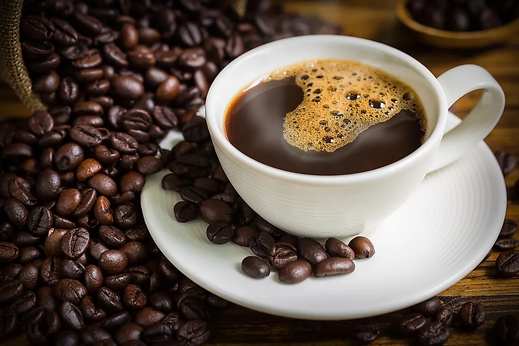 Warm close-up photo of a white cup of dark coffee with crema surrounded by scattered roasted coffee beans on a wooden surface, cozy amber-brown mood, shallow depth of field