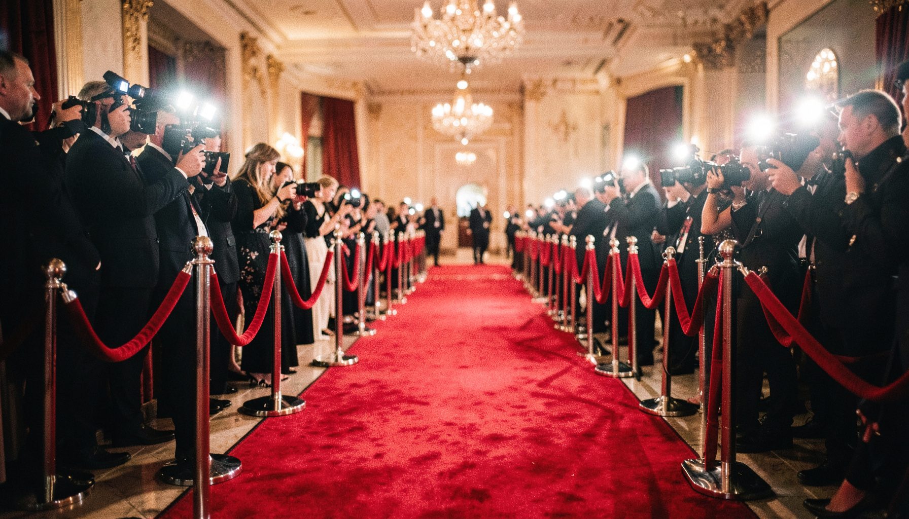 Red carpet lined with velvet ropes and camera flashes, glamorous deep red and champagne tones, celebrity-event mood
