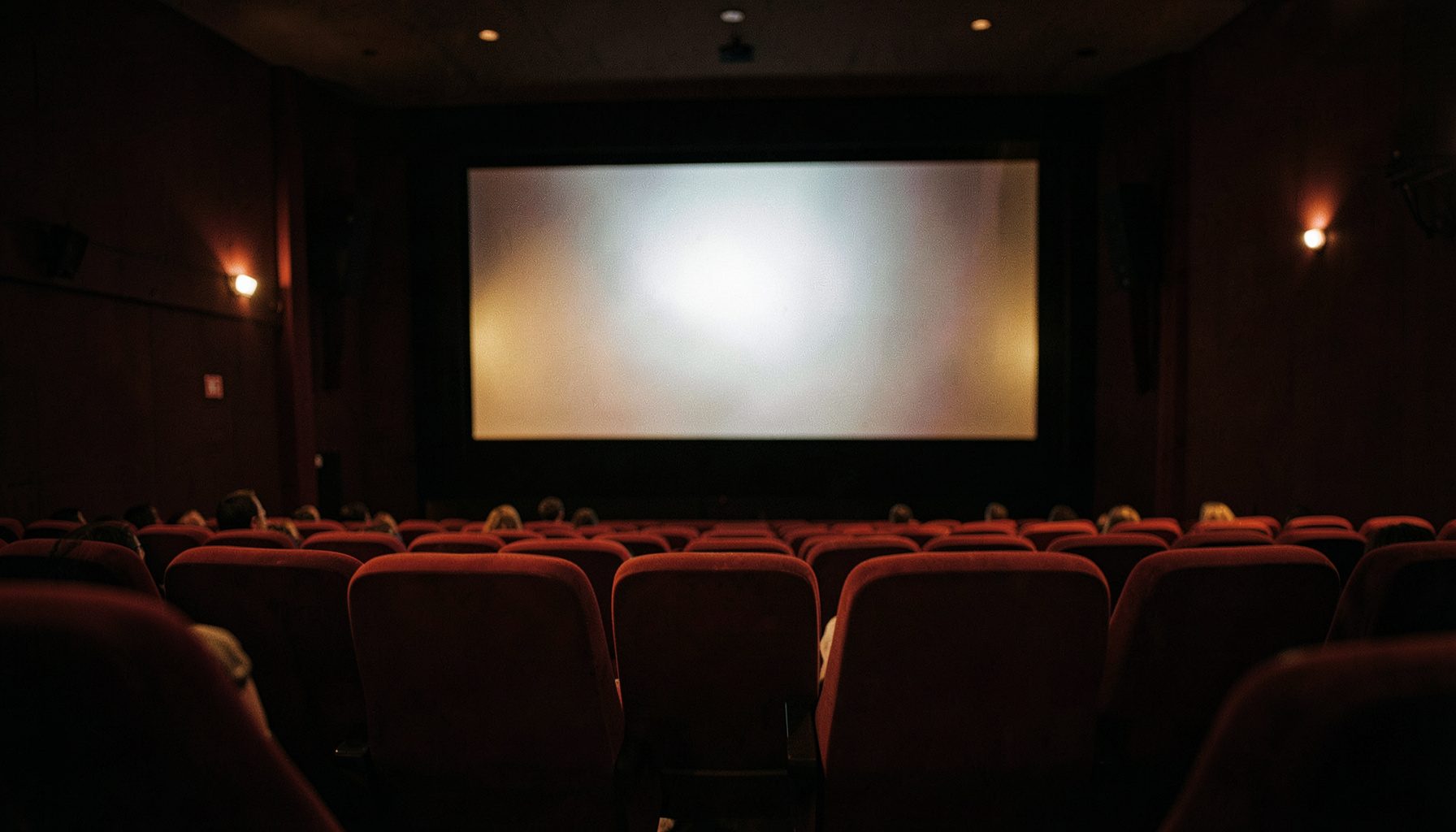 Dimly lit cinema auditorium with plush red seats and a glowing silver screen, warm amber and deep crimson tones, wide cinematic frame